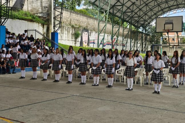 Estudiantes formadas en filas sobre la placa polideportiva cubierta, con falda a cuadros y camisa blanca; al fondo, compañeras sentadas en las gradas