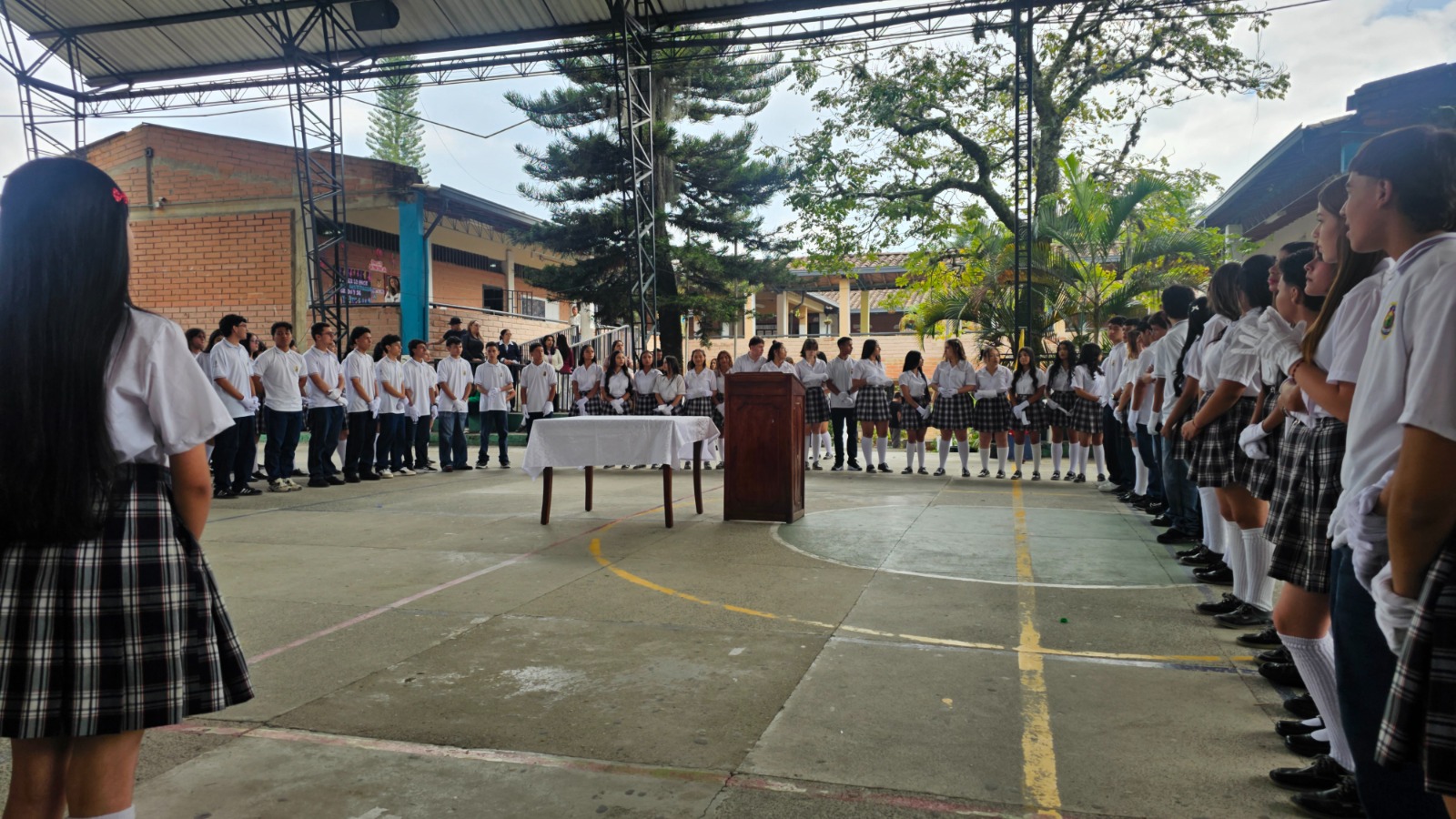 Vista general del acto cívico: estudiantes formados en círculo alrededor de un atril y una mesa en el centro de la placa polideportiva