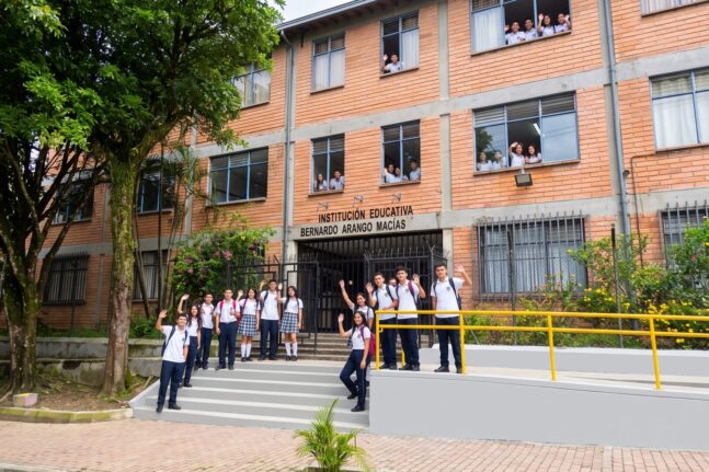 Fachada de ladrillo de la sede principal con el letrero «Institución Educativa Bernardo Arango Macías» y estudiantes de uniforme saludando en la escalera de entrada y desde las ventanas